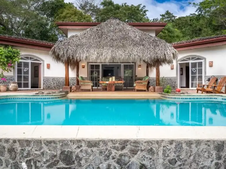 A beautiful and serene house with a distinctive thatched roof over a patio area, facing a sparkling blue swimming pool, set amidst a tropical landscape.