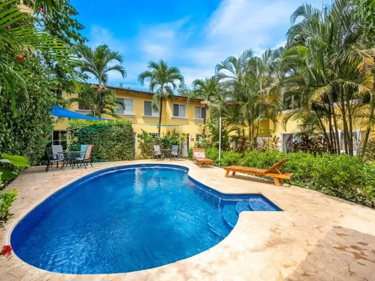 A vibrant swimming pool area surrounded by light-colored paving, lounge chairs, and lush tropical foliage including palm trees, with a yellow-toned building visible in the background under a clear blue sky.