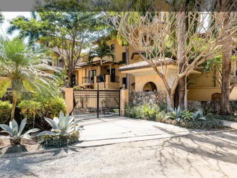 The grand entrance to a property with an ornate black gate, stone walls, and a large, multi-story building visible behind tropical trees.