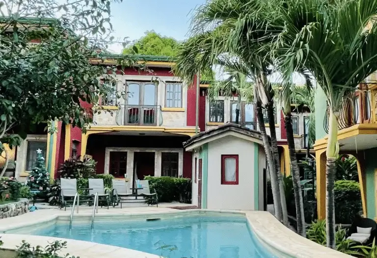 A vibrant, multi-colored building with balconies and a private swimming pool in the foreground, surrounded by lush tropical trees.