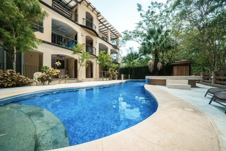 A multi-story building with balconies overlooking a curved swimming pool, surrounded by tropical trees and lush greenery.