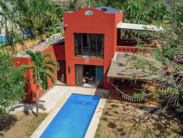 An aerial view of a striking red, multi-story modern home with a private pool, nestled amidst lush tropical greenery.