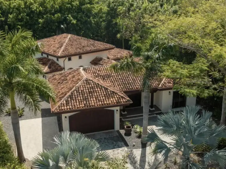 An aerial view of a beautiful, traditional-style house with a distinctive reddish-brown tiled roof, surrounded by palm trees and lush greenery.