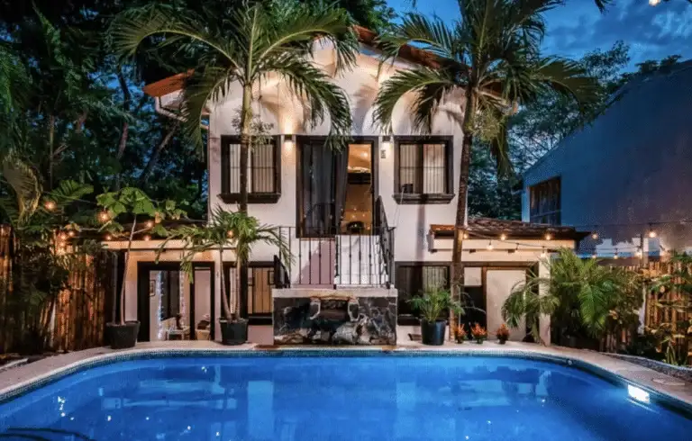 An inviting hotel exterior at dusk or night, featuring a brightly lit swimming pool in the foreground and tropical palm trees, with string lights adding ambiance.