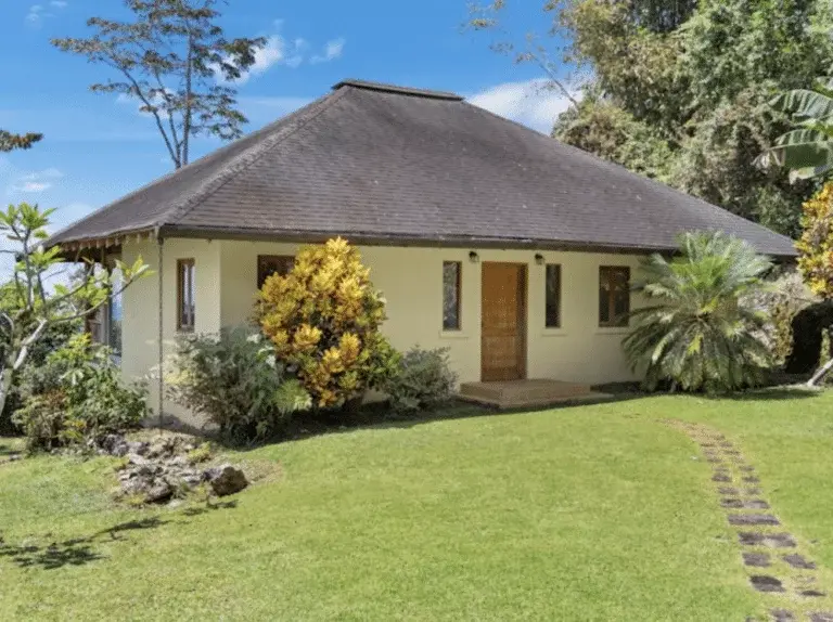 A charming single-story house with a dark roof and light-colored walls, set on a green lawn with a stone pathway, surrounded by varied tropical plants.