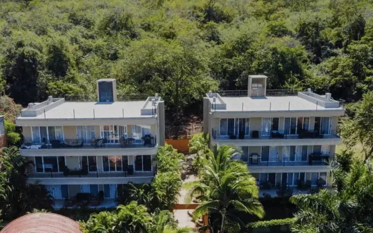 An aerial view of two multi-story, modern residential buildings with balconies, nestled amidst dense green foliage and trees, possibly near a beach.