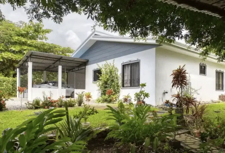 A cozy white house with a blue-grey roof and a covered carport, surrounded by a well-maintained garden with various tropical plants and a stone pathway.