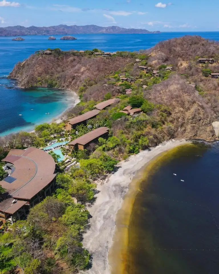 An aerial view of a stunning coastal area in Costa Rica, featuring several secluded beaches, clear blue ocean waters, and resort buildings nestled into the green and dry hillsides.