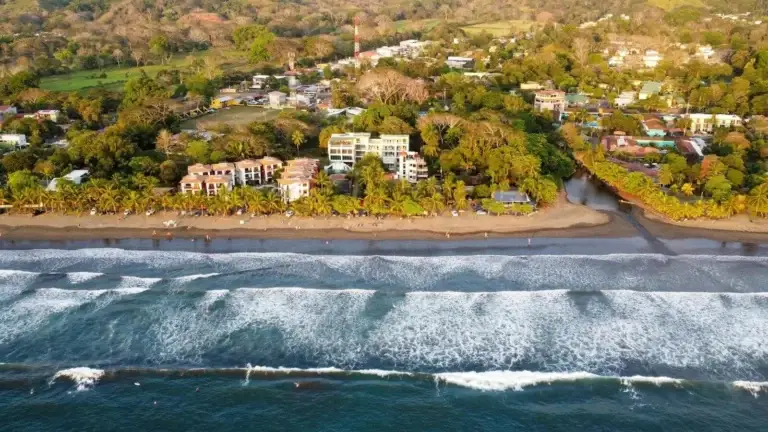 An aerial view of Jaco Beach in Costa Rica, showing the coastline with waves, a sandy beach, and a town nestled among trees and buildings along the shore.