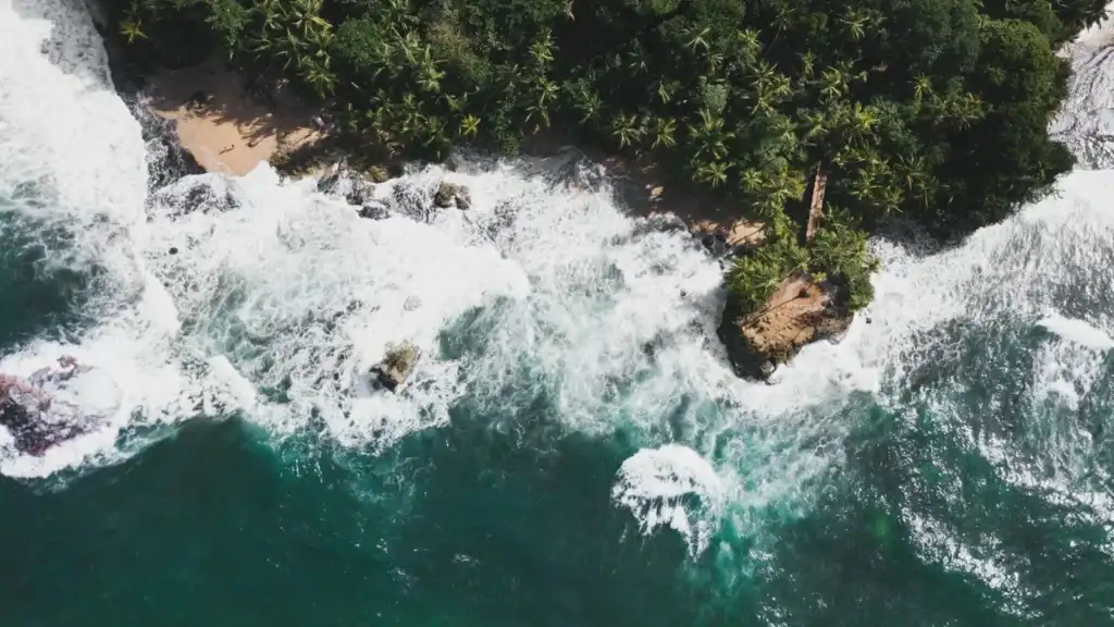 An aerial shot of a stunning tropical coastline with powerful ocean waves crashing against a sandy beach and a lush green headland.