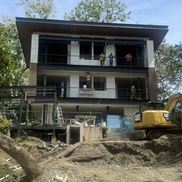 Multi-level modern villa under construction with installed roof, balcony railings, exterior cladding, and heavy equipment on site.
