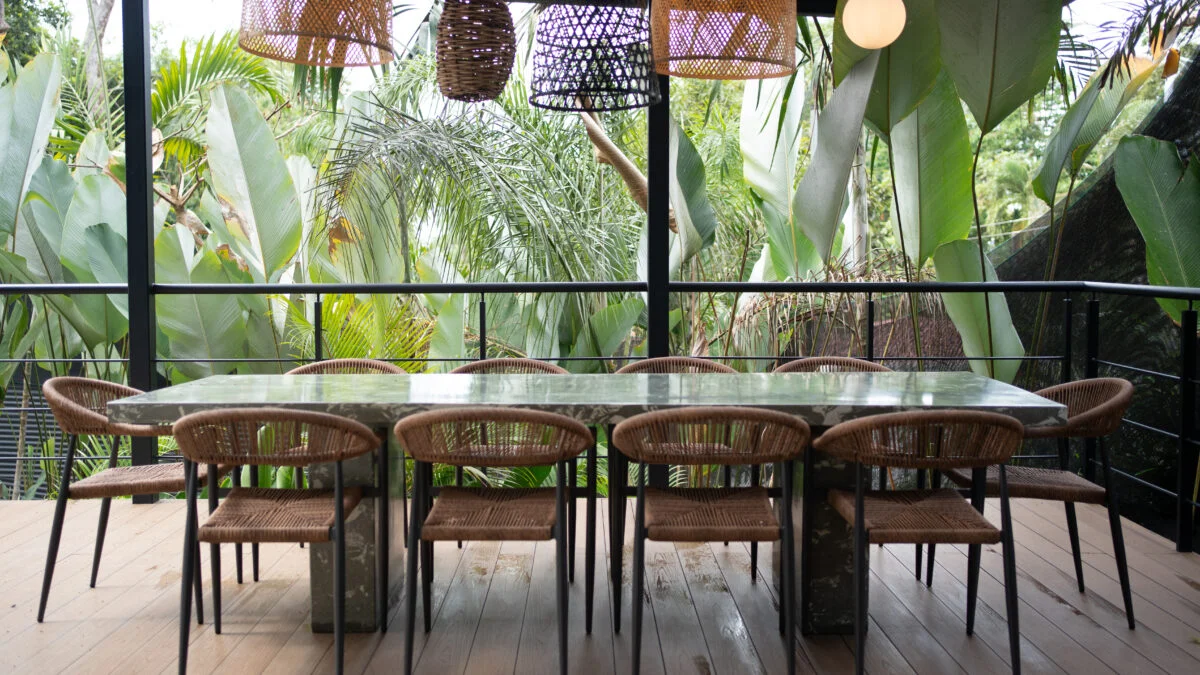 Modern jungle terrace with a concrete dining table, ten rattan-seat chairs, and unique woven hanging lamps, surrounded by dense tropical foliage.